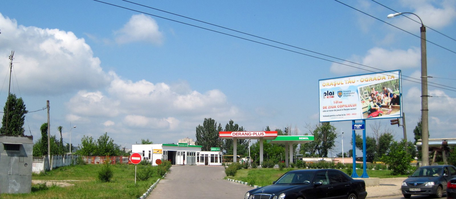 Stație de alimentare din Chișinău, cu copertina verde și roșie, o clădire albă de service auto și o mașină neagră în prim-plan
