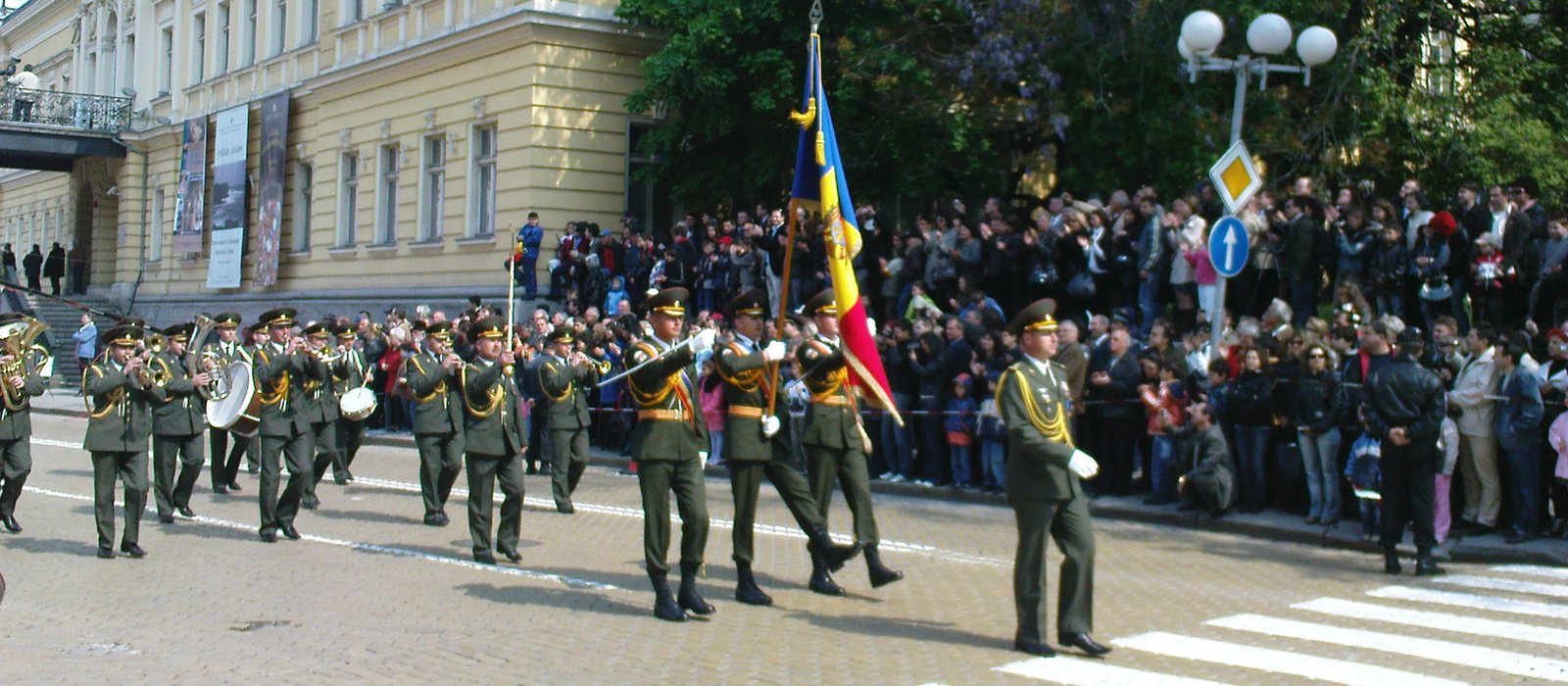 Fanfară și gardă cu drapelul tricolor al Republicii Moldova defilează pe o stradă, cu spectatori de-a lungul trotuarului