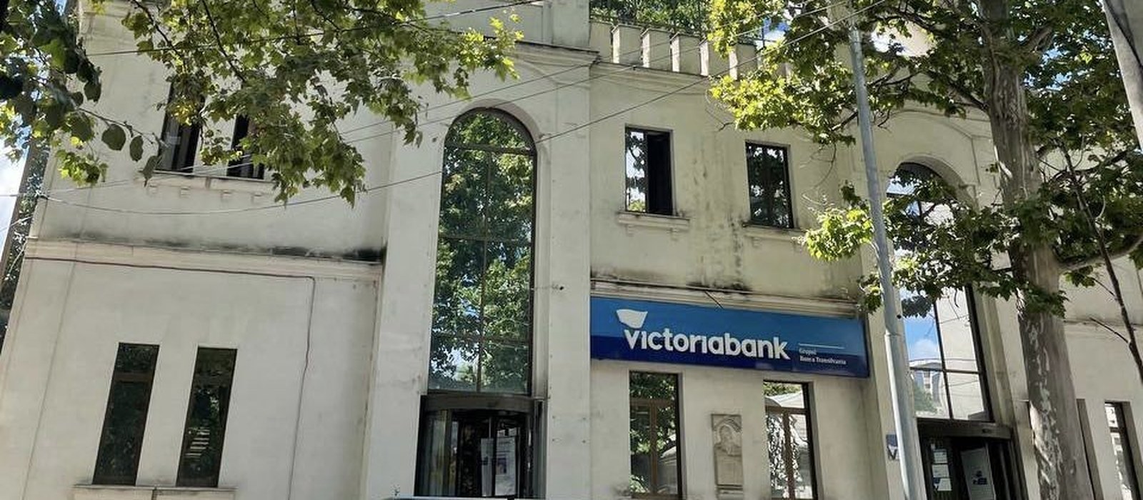 Sediul Victoriabank din Chișinău, clădire albă cu fațadă înaltă, cu panoul albastru al băncii la parter, printre crengi de platan