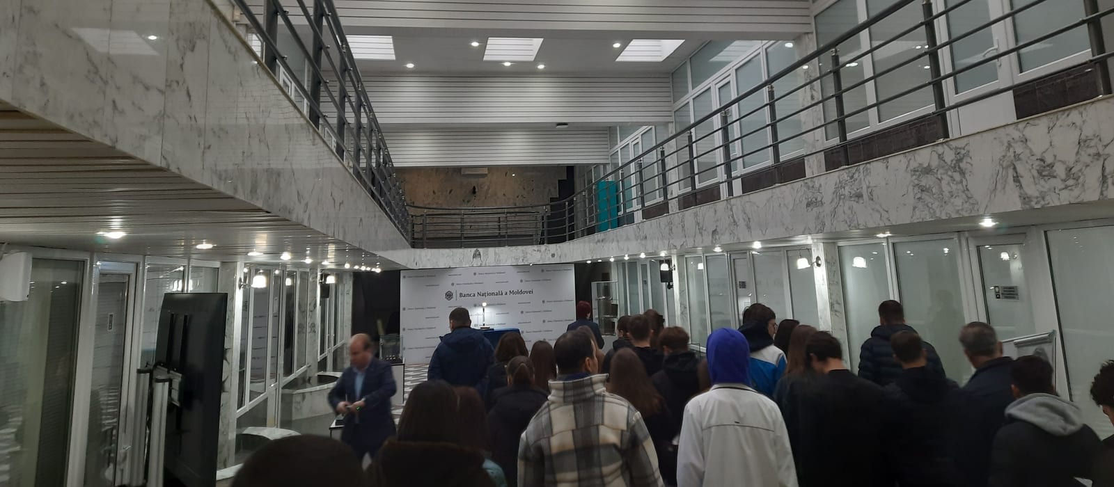 Holul Băncii Naționale a Moldovei, cu marmură albă, balustrade metalice și un grup de vizitatori în fața panoului cu sigla băncii