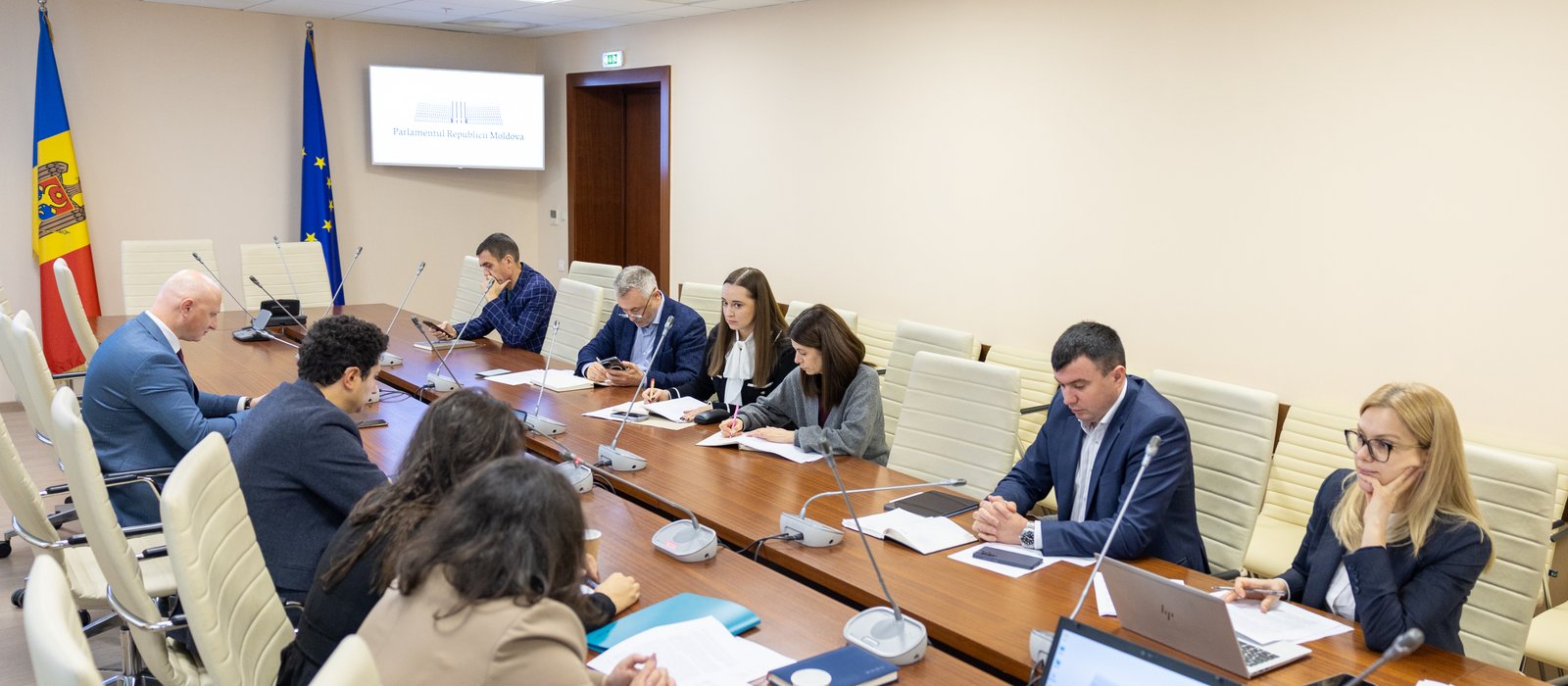 Sală de ședințe a Parlamentului Republicii Moldova, cu steagurile Moldovei și Uniunii Europene și oameni la masă cu documente