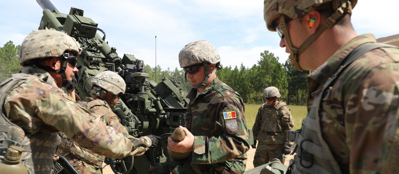Un militar al Armatei Naționale a Moldovei ține un proiectil de artilerie lângă un obuzier, între militari americani, pe un poligon cu pini