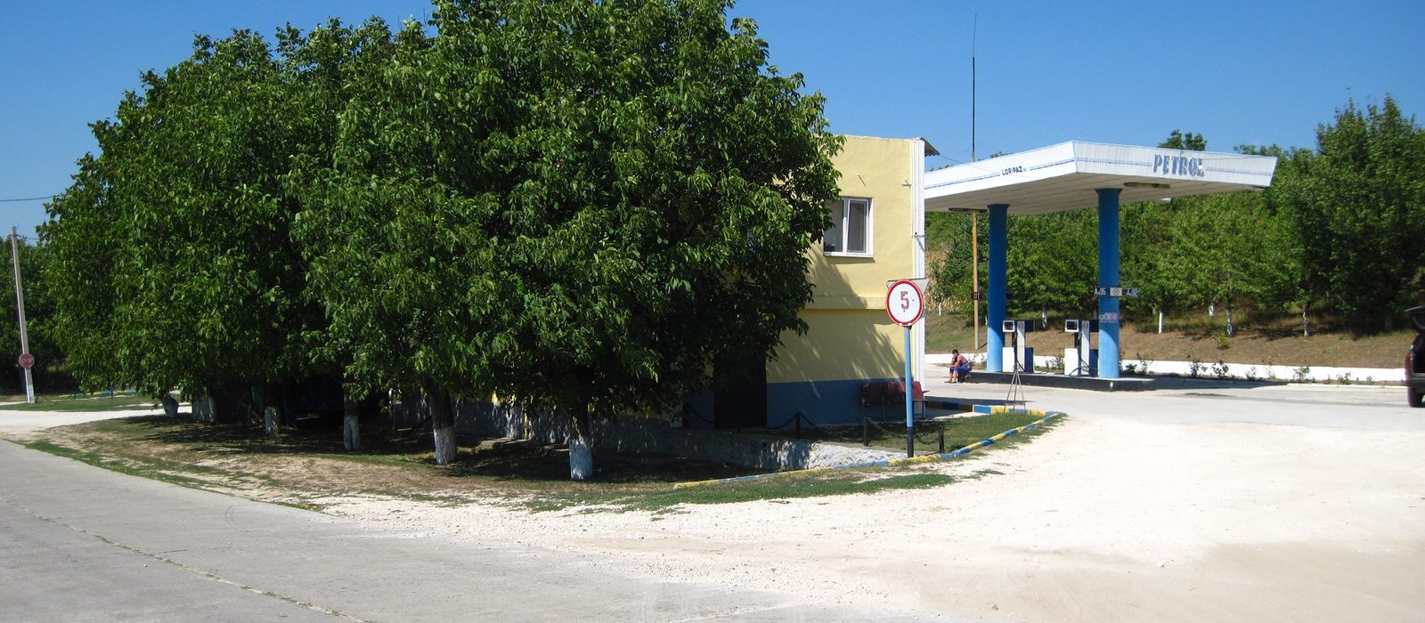 O stație de alimentare cu două pompe sub un acoperiș albastru, lângă o clădire galbenă și un nuc mare, într-un sat din raionul Strășeni