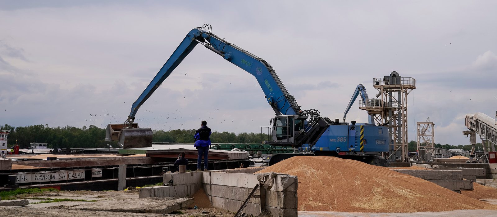 Un excavator cu graifăr încarcă grâu dintr-un morman pe o barjă, într-un port dunărean, sub un cer noros
