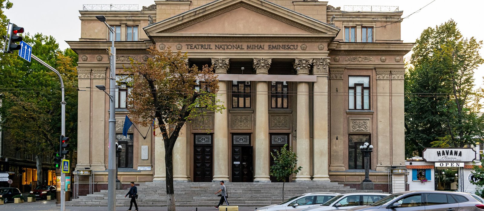 Fațada cu coloane a Teatrului Național Mihai Eminescu din Chișinău, cu doi trecători pe treptele din fața clădirii