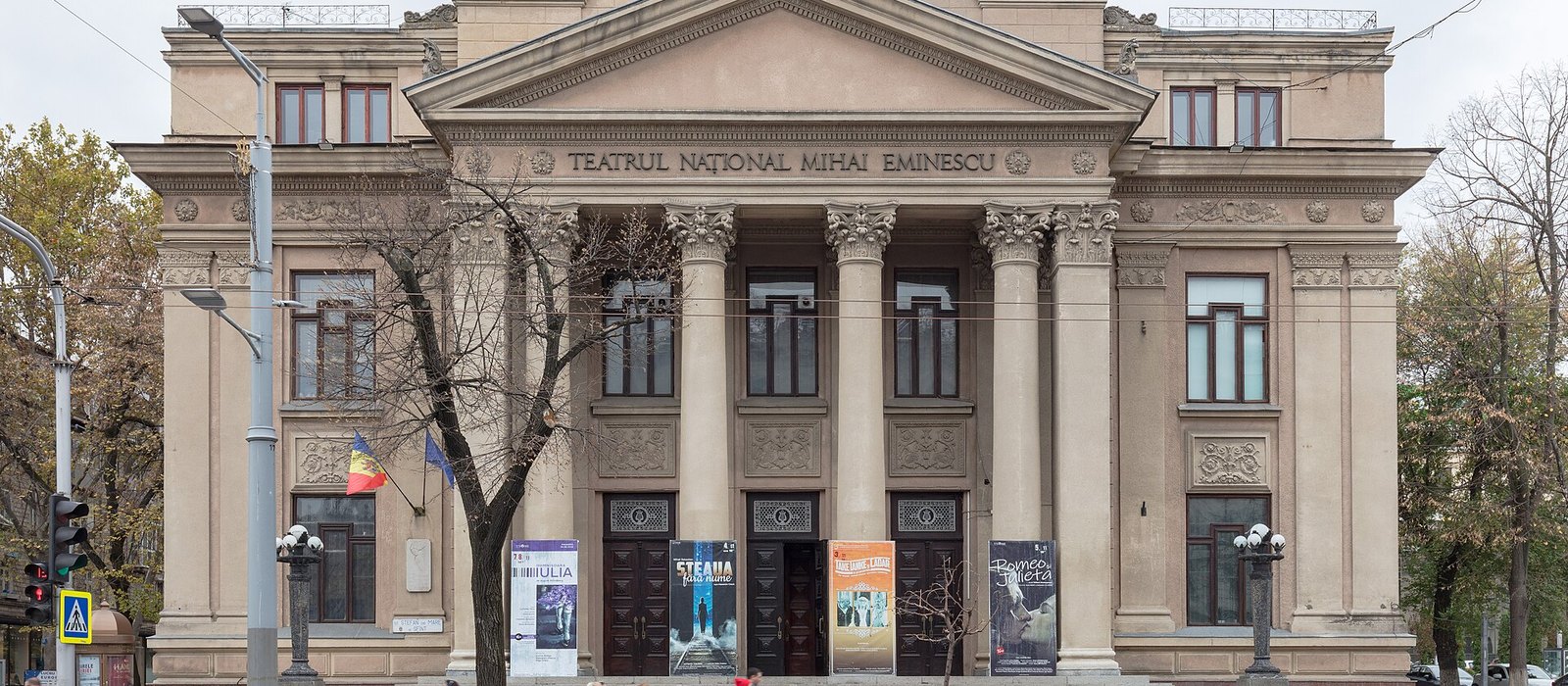 Fațada cu coloane a Teatrului Național Mihai Eminescu din Chișinău, cu afișe pe ușile de la intrare și drapelele Moldovei și UE