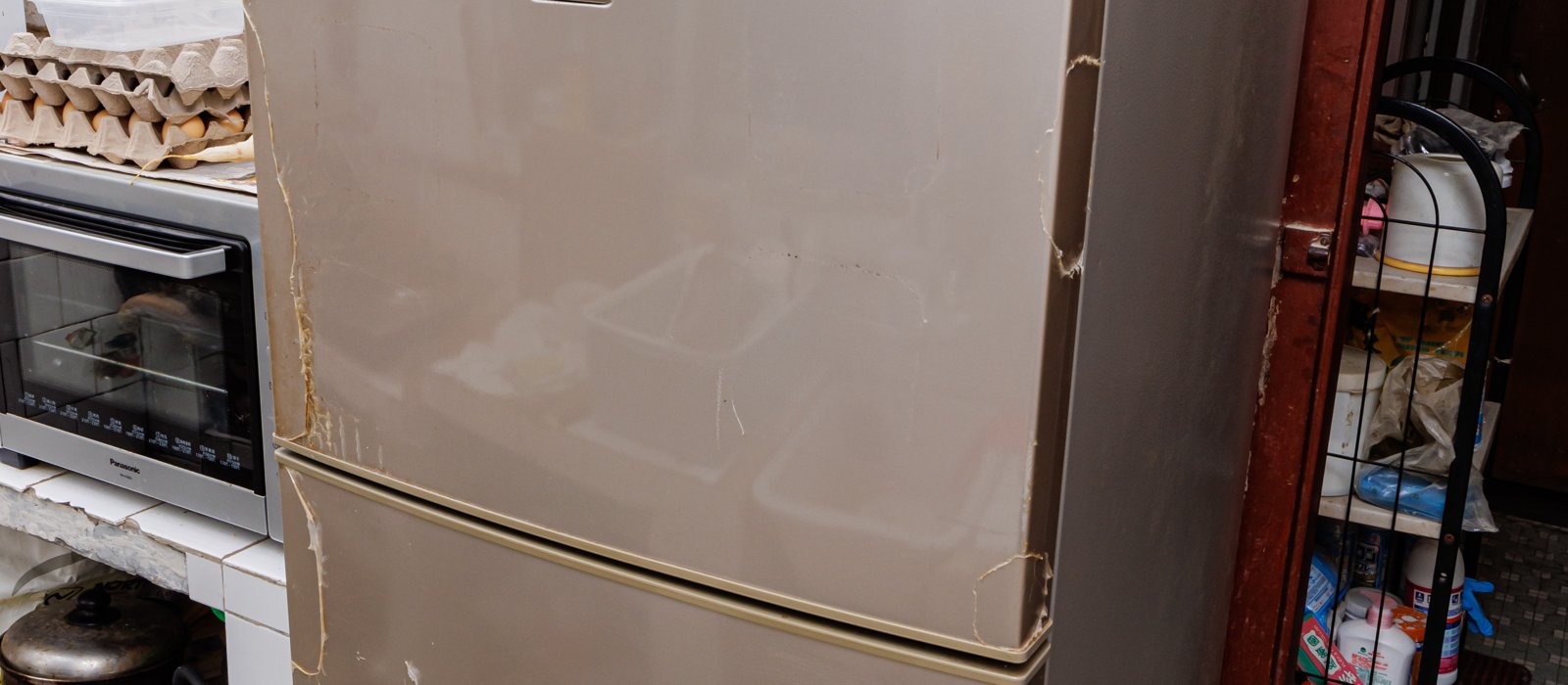 Un frigider vechi, cu colțurile uzate și mâzgălit pe uși, într-o bucătărie, lângă un cuptor electric