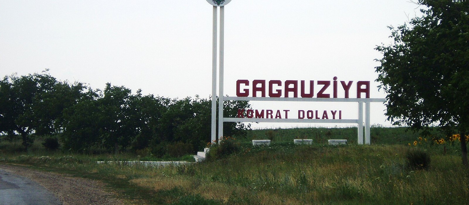 Panou alb cu stema și numele Găgăuziei la marginea unui drum, lângă o plantație