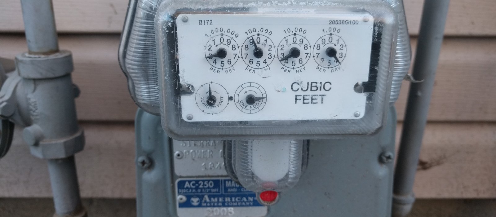 Contor de gaz natural cu patru cadrane, montat pe peretele exterior al unei case, între două țevi