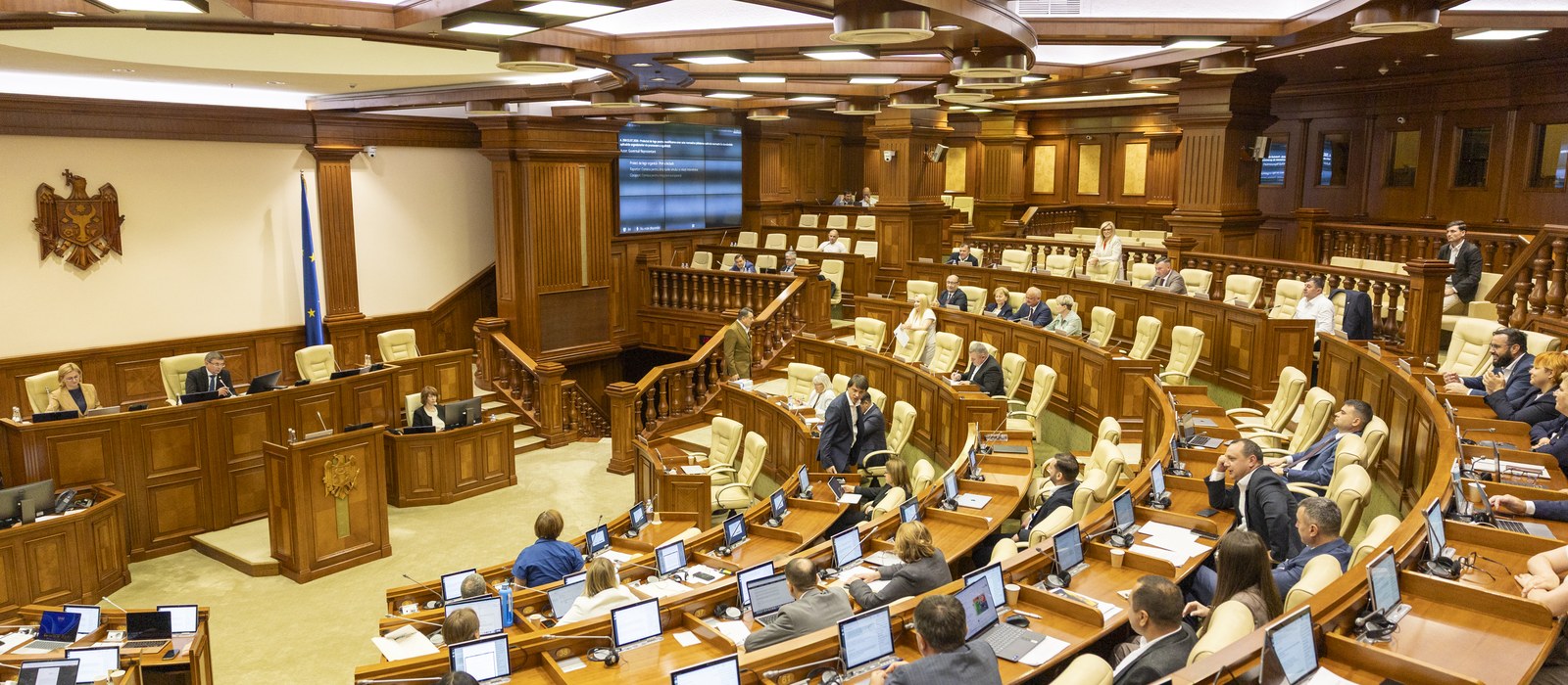 Sala de plen a Parlamentului Republicii Moldova în timpul unei ședințe, cu deputați la bănci și stema de stat pe peretele din stânga