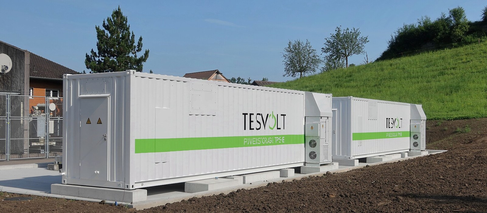 Două containere albe de baterii ale unei stații de stocare a energiei, pe un teren deschis