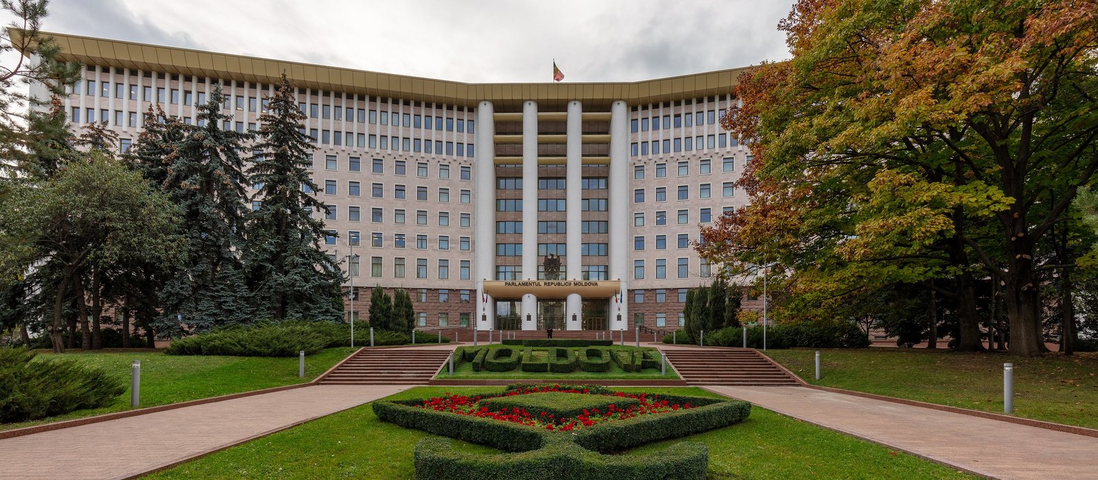 Fațada Parlamentului Republicii Moldova la Chișinău, cu un rond de flori roșii și inscripția MOLDOVA din tuiuri în față și arbori în culorile toamnei