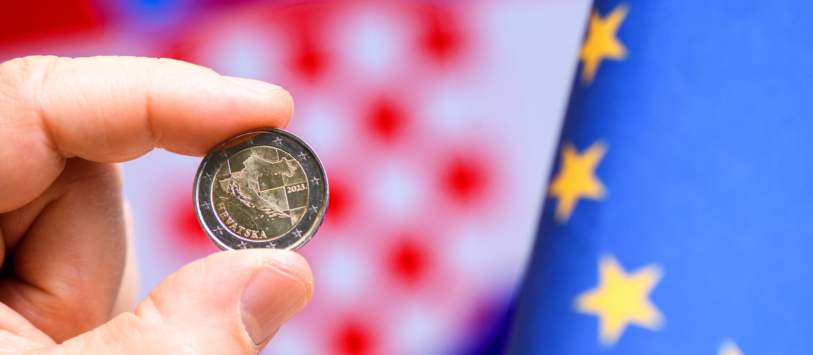 O mână ține între degete o monedă de 2 euro croată, în fața unui steag albastru cu stele galbene ale Uniunii Europene