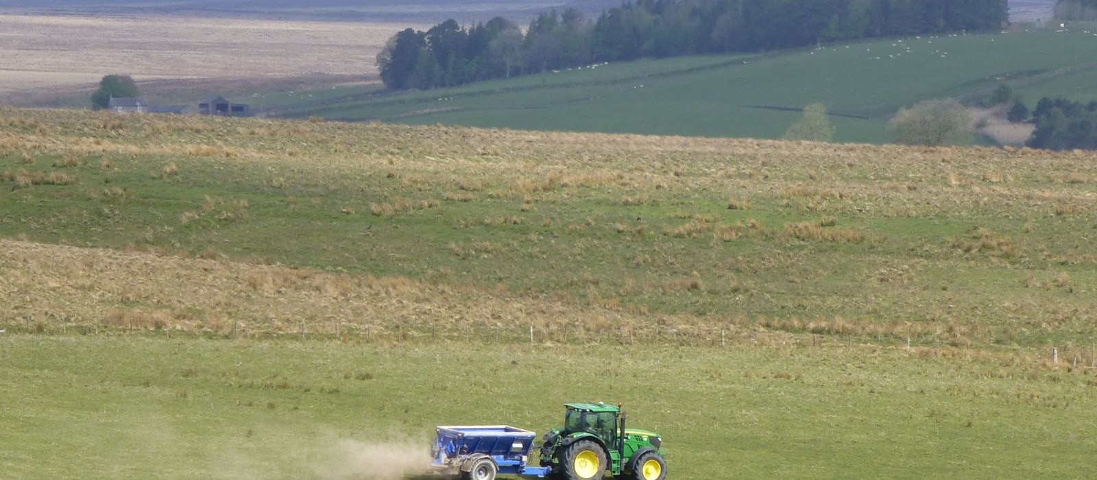 Tractor verde cu remorcă albastră care împrăștie îngrășământ pe un câmp, cu dealuri în fundal
