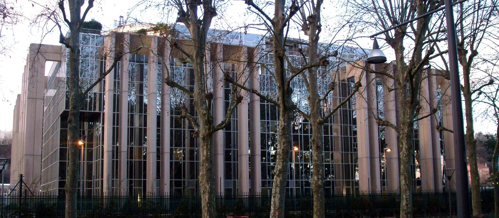 Fațada de sticlă și coloane de piatră a sediului Interpol din Lyon, văzută printre copaci iarna