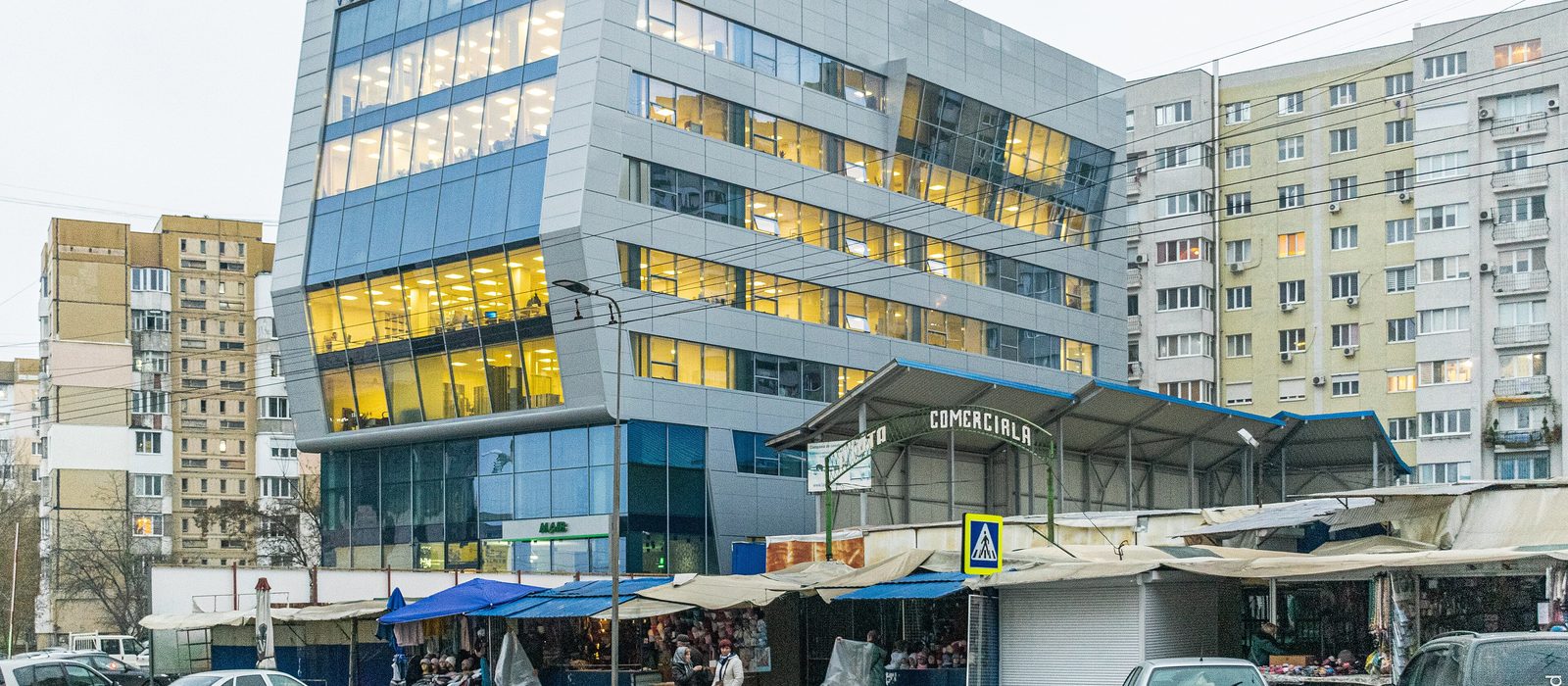 Clădire de birouri cu etajele luminate, între blocuri de locuințe și piața acoperită dintr-un cartier al Chișinăului