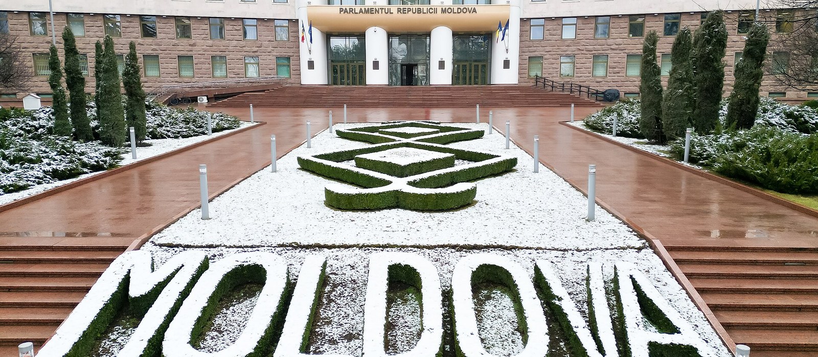 Fațada Parlamentului Republicii Moldova cu denumirea instituției deasupra intrării și inscripția MOLDOVA din arbuști tunși în fața scărilor