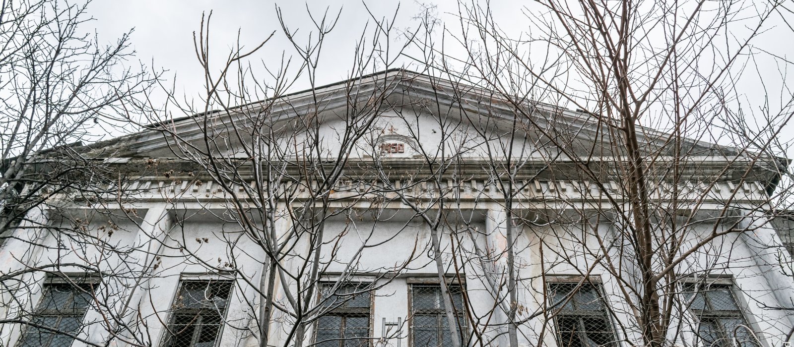Fațada unei clădiri industriale vechi din Chișinău, văzută printre crengi fără frunze, cu anul 1950 în fronton