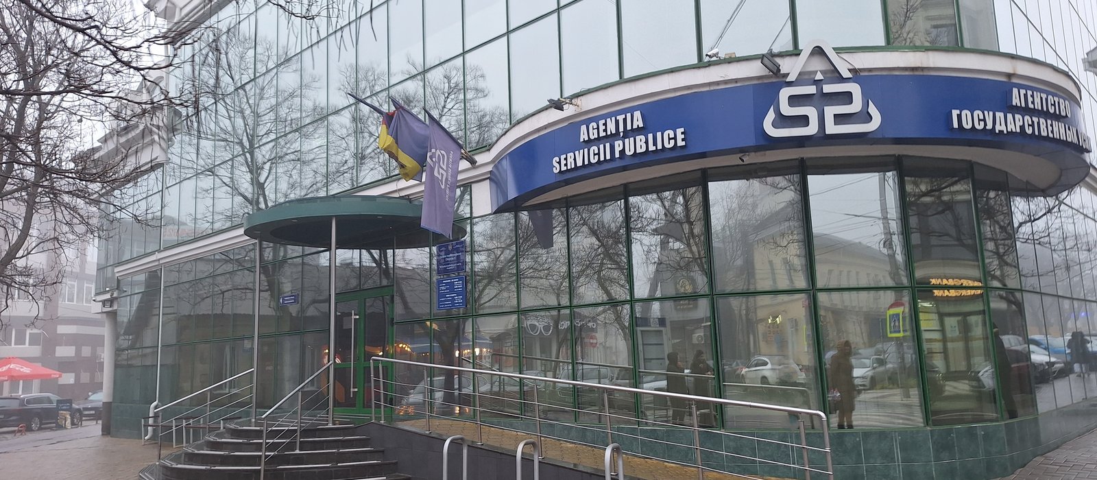 Clădire din sticlă cu fațadă curbă și panou albastru la intrare, într-o zi cu ceață, pe o stradă din centrul Chișinăului