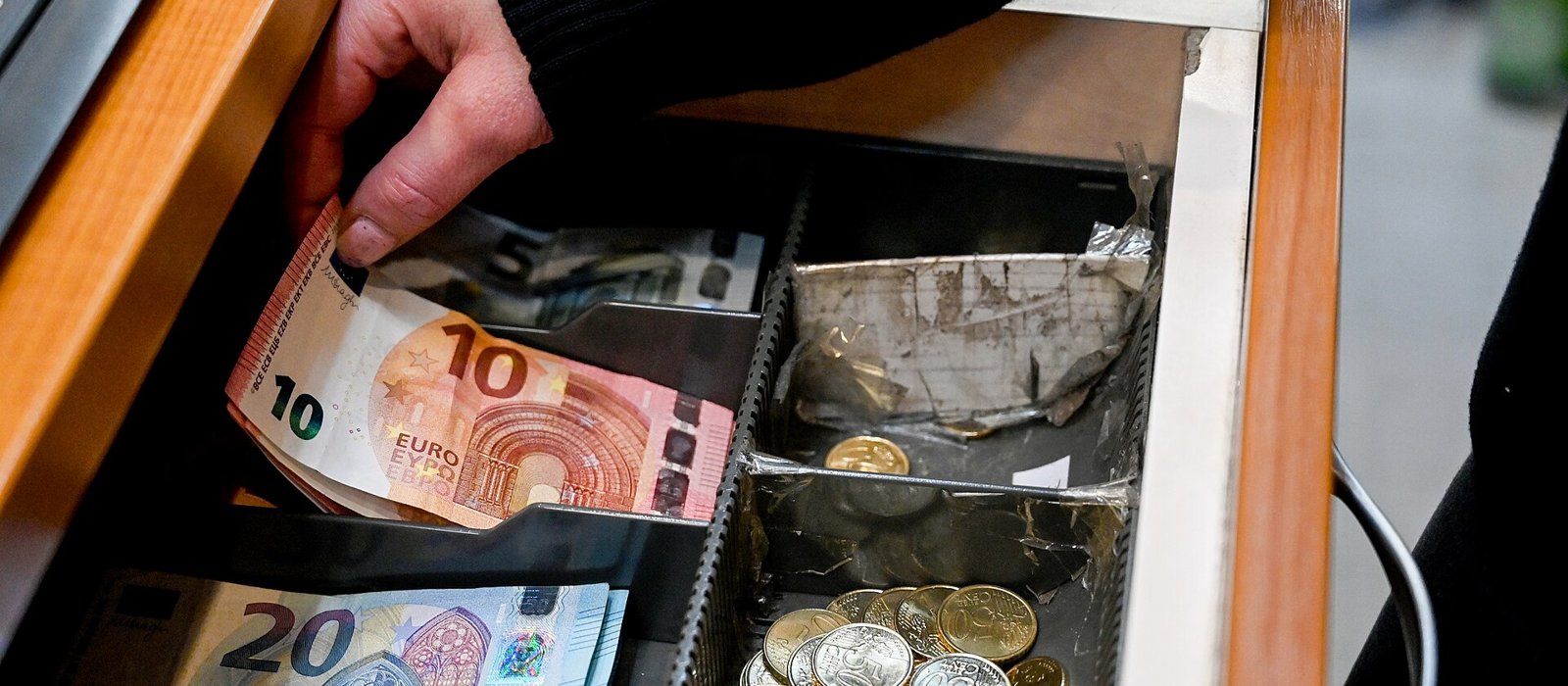 O mână scoate o bancnotă de 10 euro dintr-un sertar de casă, lângă monede și o bancnotă de 20 de euro